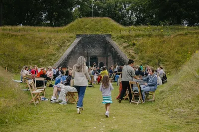Bezoekers zitten op stoelen en kleedjes in het gras voor De Poterne